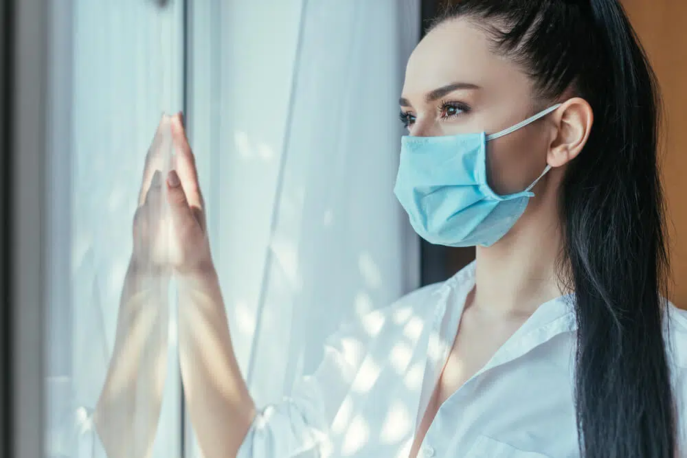 Thoughtful girl in medical mask looking through window at home