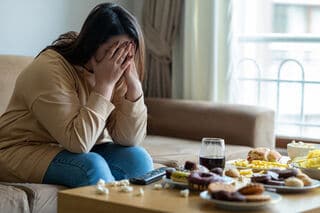 Woman struggling with binge eating covers face with lots of food spread on table before her.