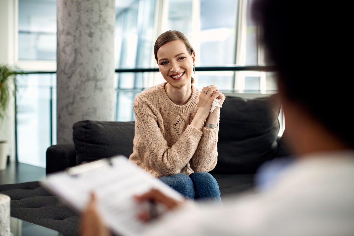 A woman smiling during a therapy session while the therapist holds a clipboard.