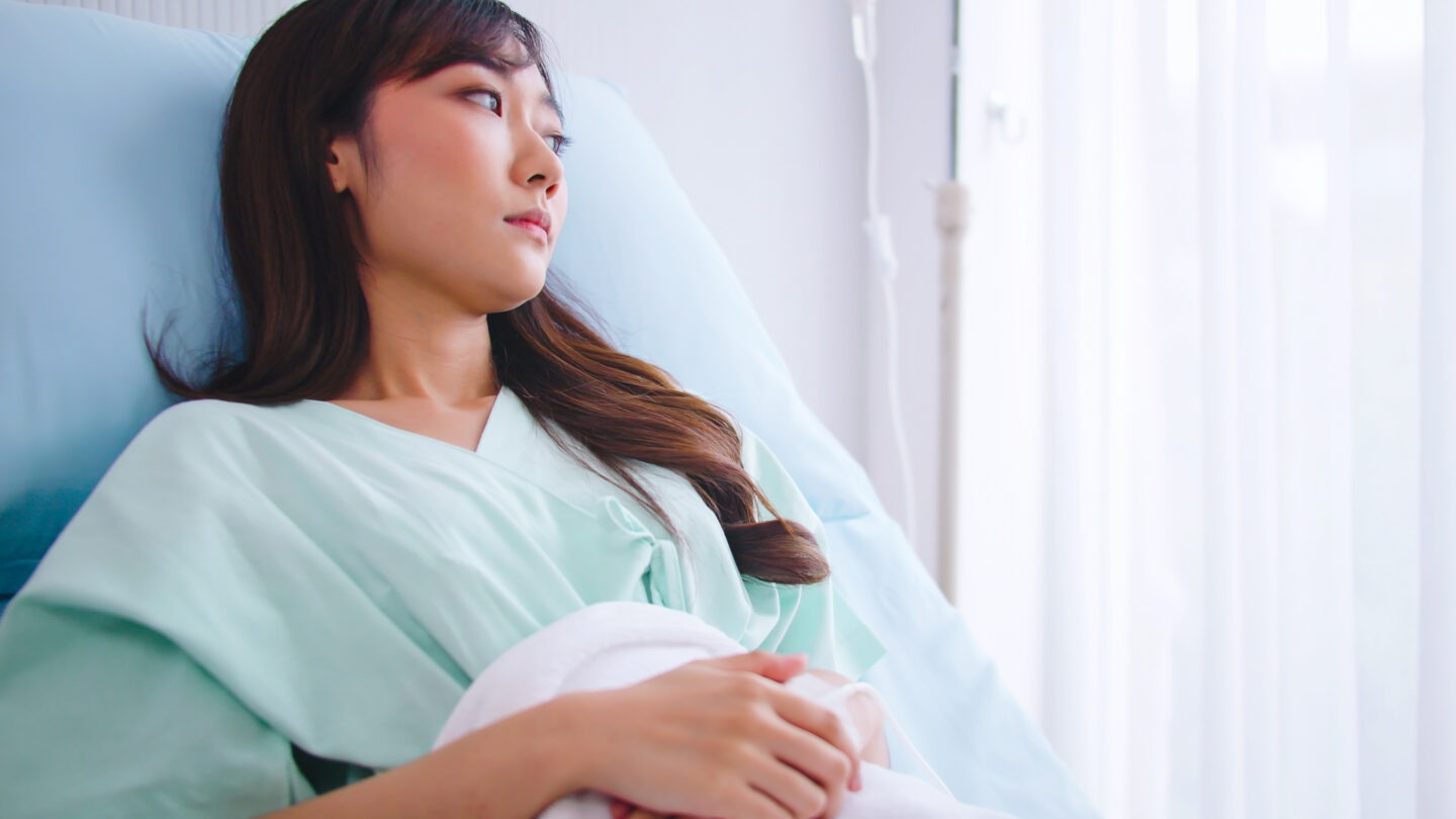 A sadlooking woman looking out of a window while laying in a hospital bed.